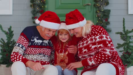 Elderly-grandmother-grandfather-with-granddaughter-sitting-at-Christmas-house-porch-waving-hello-hi