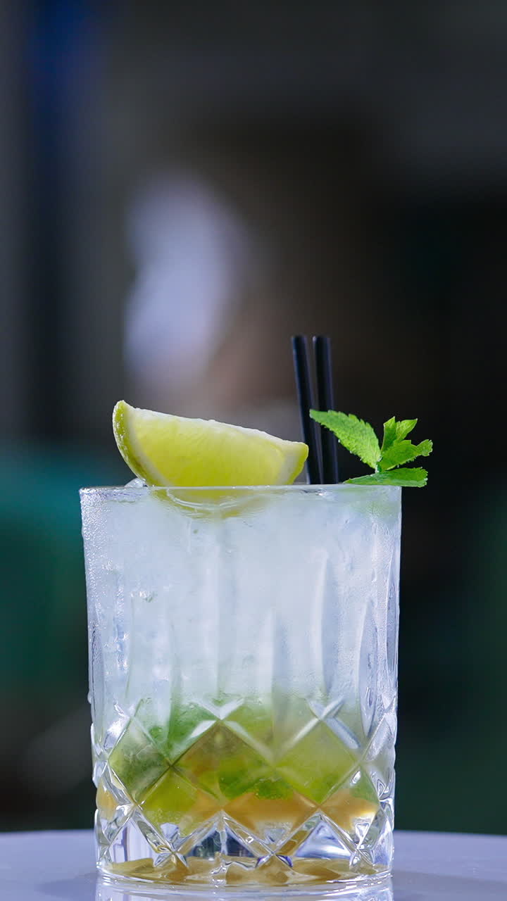 Beverage in old-fashioned glass decorated with lemon and mint leaves. Cocktail with two straws on the rotating table. Blurred backdrop. Close up. Vertical video