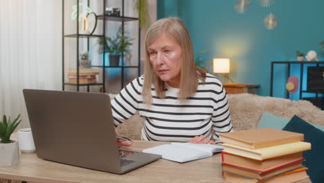 mature woman participating in online course on laptop making notes sits at table with books nearby