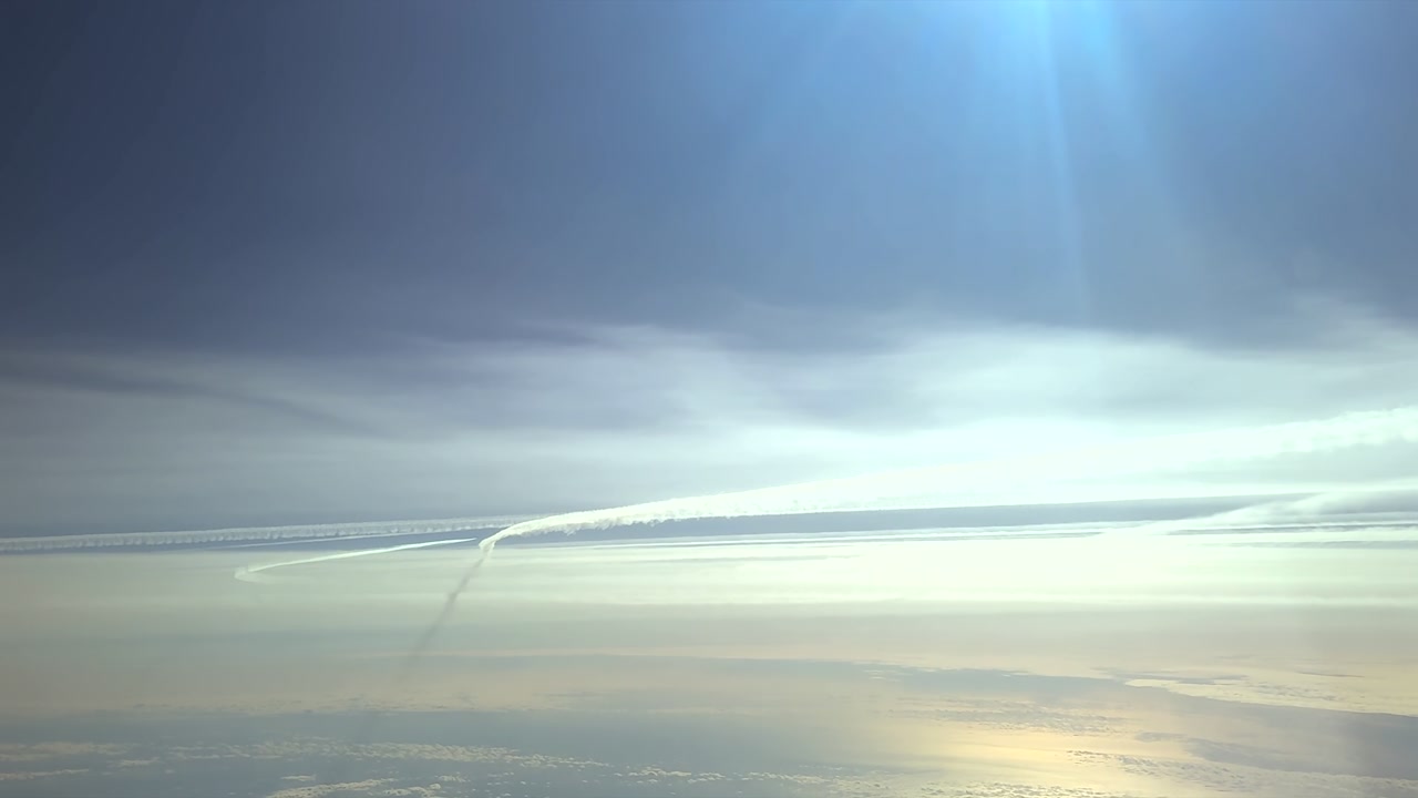 pilot view from a jet cockpit flying in the upper atmosphere, with a view of multiple contrails in a blue sky, with a radiant sun shining above the sea.