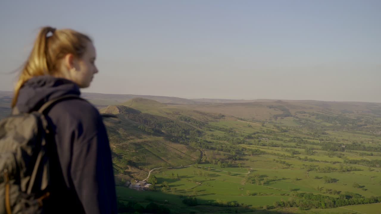Ripresa a mano, panoramica rotante di una giovane donna bionda con i capelli raccolti e uno zaino che ammira il panorama da Mam Tor, Castleton, Peak District, Inghilterra.