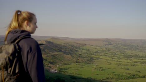 Handheld-rotating,-panning-shot-of-young-blonde-woman-with-hair-tied-up-and-backpack-admiring-the-view-from-Mam-Tor,-Castleton,-Peak-District,-England