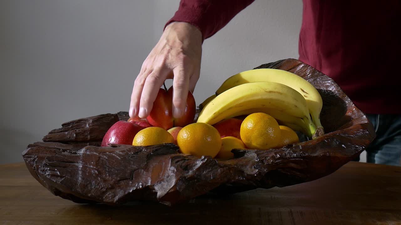 una mano masculina toma una manzana de un tazón de madera con frutas sobre una mesa de madera