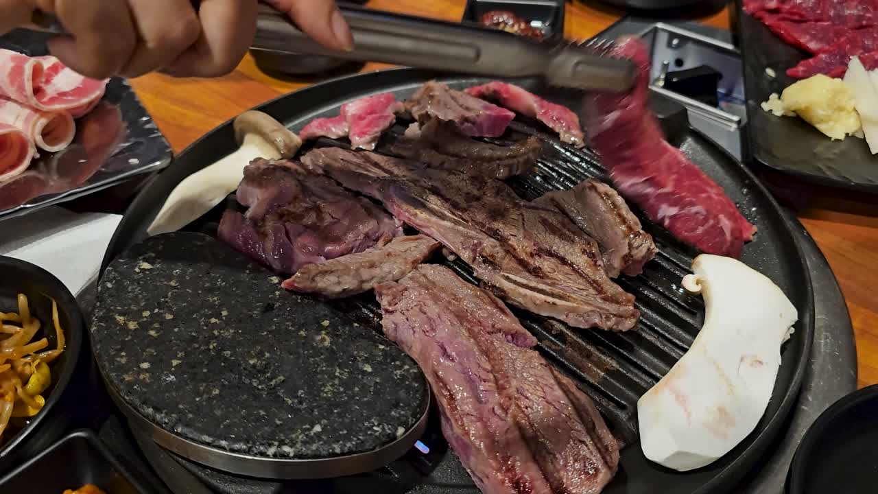 Close up of delicious beef cuts like galbi grilling on a traditional Korean barbecue, with tongs turning the sizzling meat alongside mushrooms and side dishes