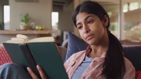 Video-of-biracial-woman-sitting-on-sofa-and-reading-book