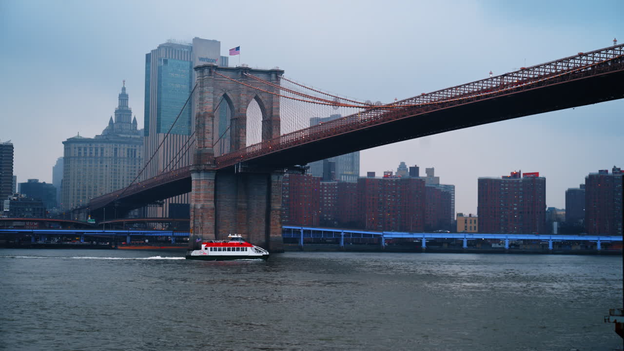 New York, USA, 25 dicembre 2025: Un traghetto NYC passa sotto il Ponte di Brooklyn a Lower Manhattan. Sull'East River, un'imbarcazione rossa e bianca con il Ponte di Brooklyn.