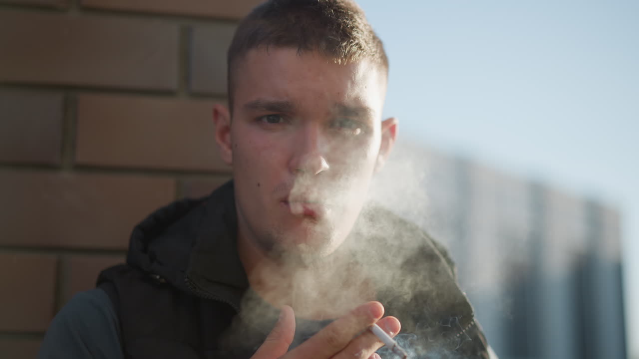young man standing outdoors against brick wall exhales cigarette smoke slowly with focused, contemplative expression, sunlight hitting face while soft urban background fades into distance