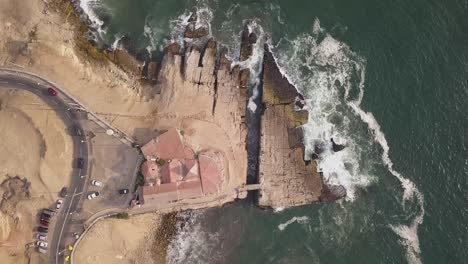 dynamic aerial shot capturing ocean waves crashing against rugged cliffs, featuring a unique coastal building. ideal for travel, real estate, or high-resolution stock imagery. leap of the friar.