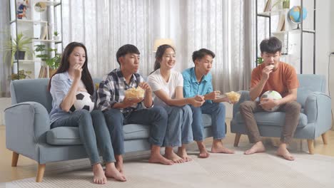 asian teenagers cheering, eating and watching football game on tv at home