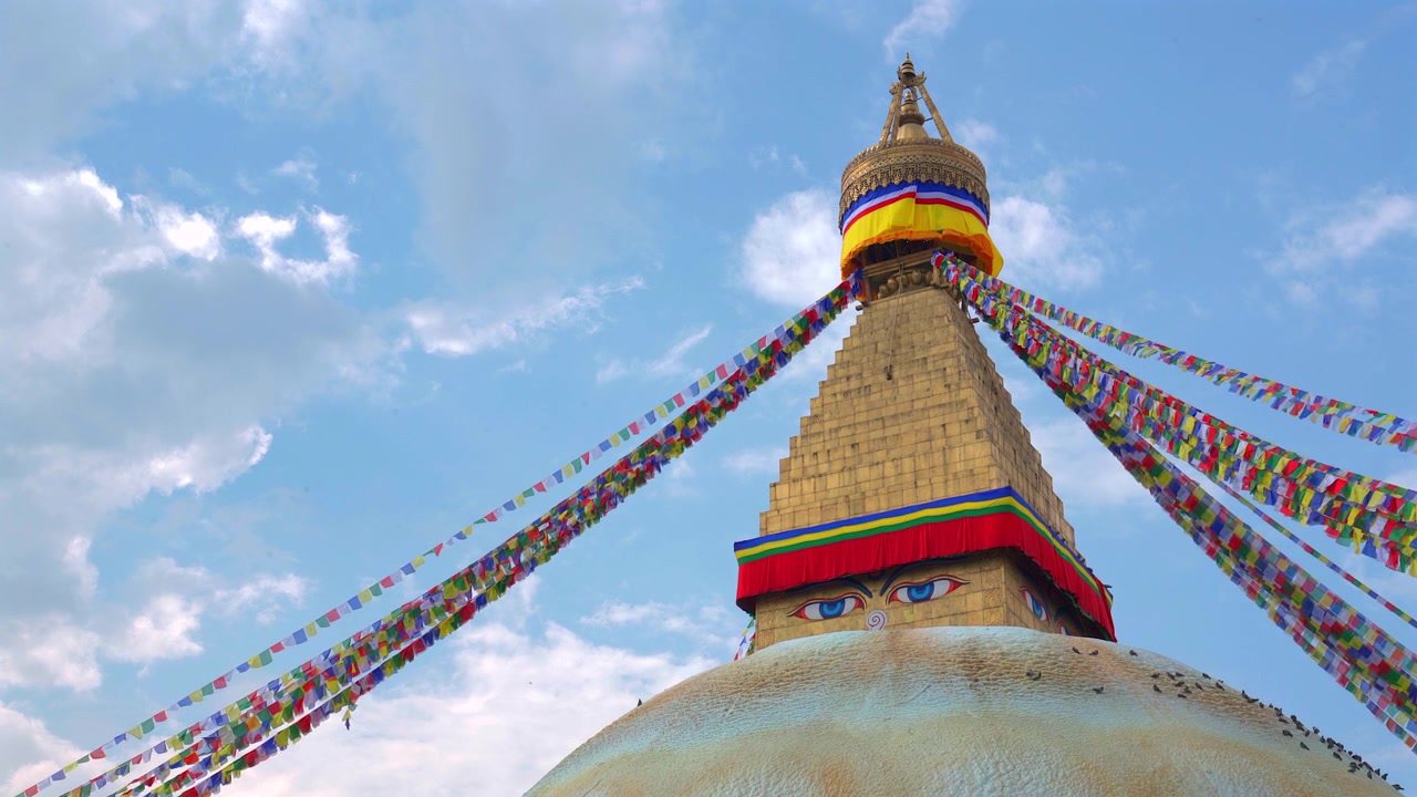 Ripresa statica dal basso dell'iconica guglia dorata e degli occhi della saggezza della Stupa di Boudhanath, adornata con vivaci bandiere di preghiera buddiste che si estendono sotto un cielo blu con soffici nuvole bianche a Kathmandu.