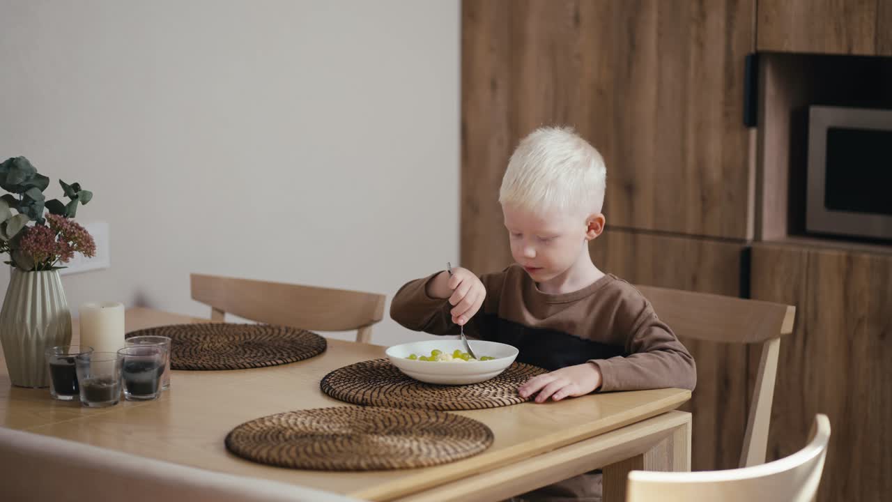 Little albino boy with white hair eats oatmeal with grapes while having breakfast while sitting on a wooden chair in a modern kitchen. Little albino boy with white hair has breakfast in the kitchen