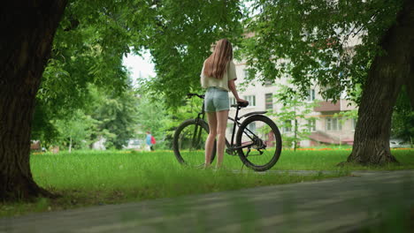 young woman in pink sneakers approaches her bicycle on tree-lined path, placing an item on seat, background features lush greenery, a building, and faint silhouette of a person in distance