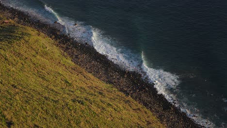 Rolling-waves-breaking-on-rock-shore-with-grass-slope-cliff,-slow-motion