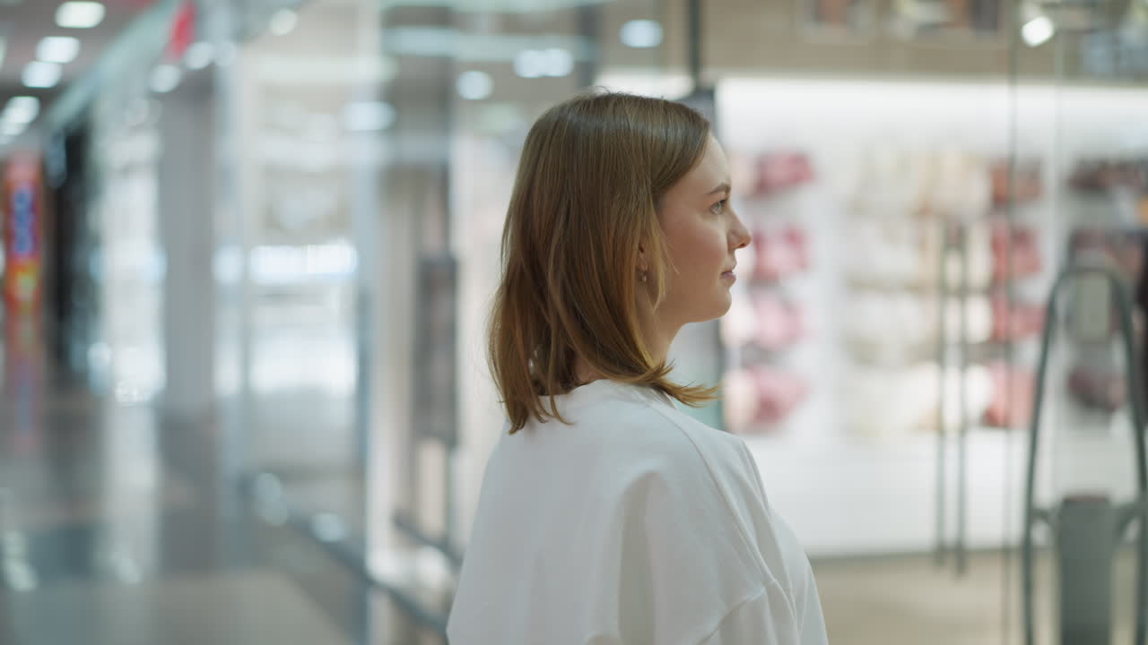 Lady stands in front of cloth store in mall observing mannequin dressed in pants and bra, with blur background featuring racks of clothing, handbags, and bright retail displays in a well-lit mall