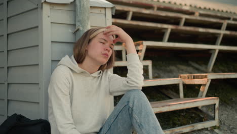 lady in jeans seated on ground with hand resting on head in thoughtful expression, situated in a stadium with worn bleachers in background, black bag beside her