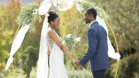 african american couple holding hands and smiling at their wedding in sunny garden, in slow motion