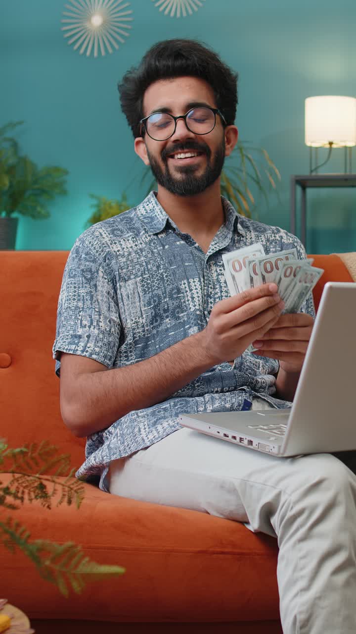 Smiling indian man counting money cash and use laptop pc calculate domestic income earnings at home