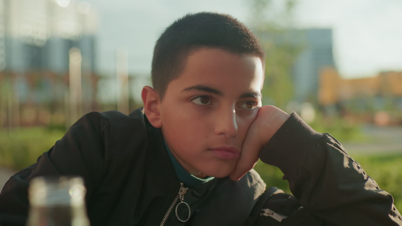 Close up of boy thoughtfully sketching on notebook with head resting on hand while adult offers chips and juice on wooden table outdoors, calm study moment with snacks in focus