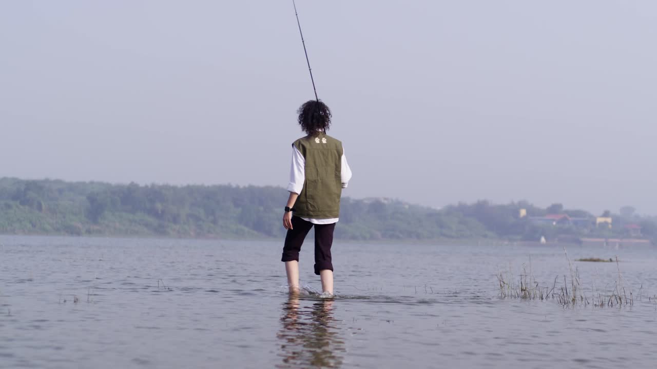 Seorang gadis Asia Selatan mengenakan rompi memancing berjalan dengan percaya diri melalui air danau sambil memegang pancingnya