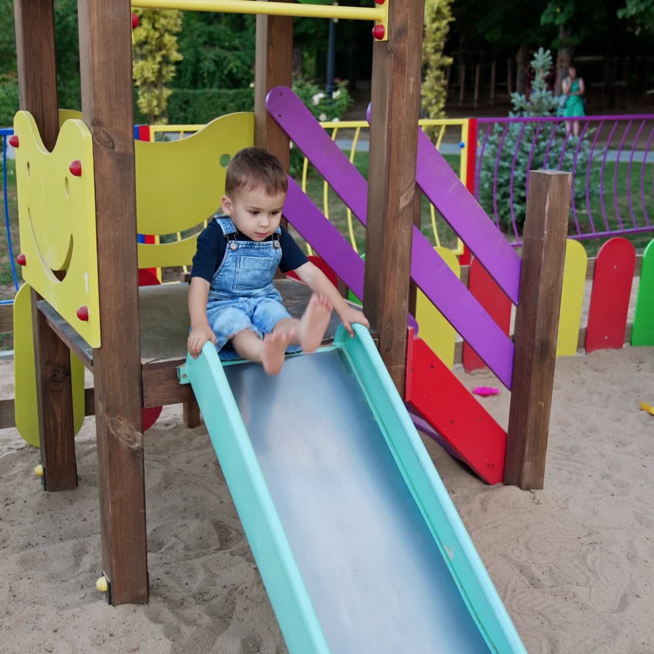 Lovely baby banging feet by the slide on the playground. Kid in a jeans romper goes down by the slide