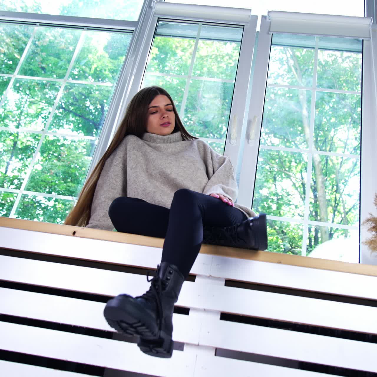 Sad beautiful girl with long dark hair wearing warm sweater sitting on the window-sill. Low angle view. Green trees in window at backdrop
