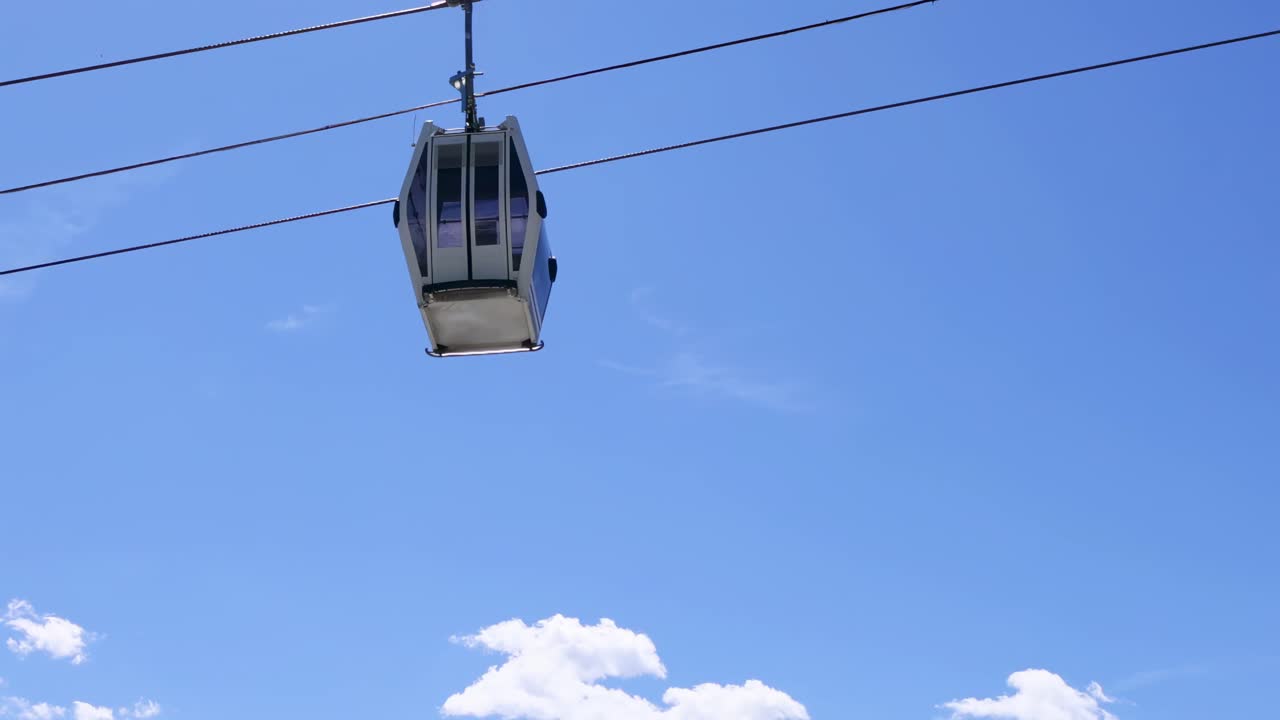 A cable car glides above a lush forest with majestic mountains in the background under a clear blue sky.