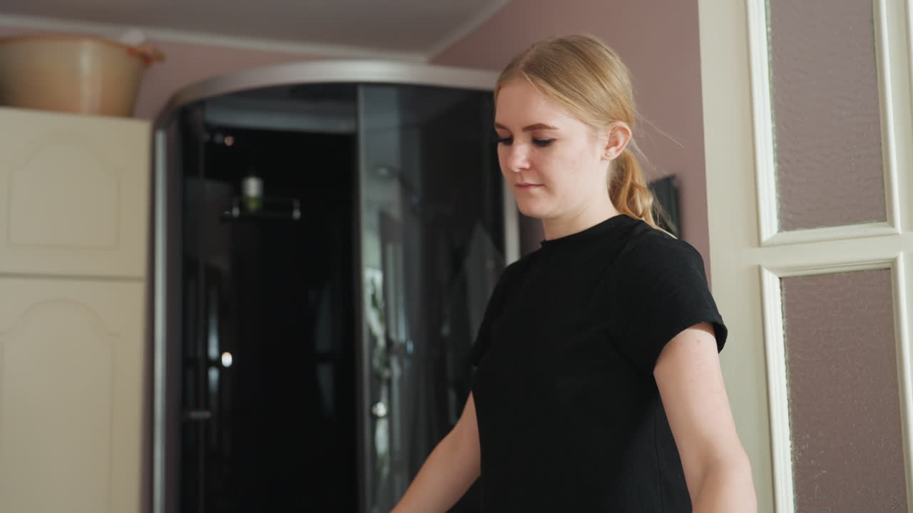 Blond woman wearing silver earring holds white bed sheet while tidying bed in softly lit room, with subtle reflection visible in wall mirror and gentle blur in background highlighting calm atmosphere
