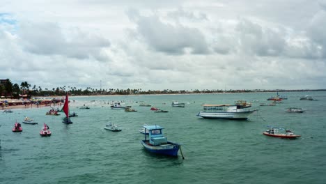 Slow-dolly-in-aerial-drone-shot-of-the-Porto-de-Galinhas-or-Chicken-Port-beach-with-anchored-sailboats-and-tourists-swimming-in-the-crystal-clear-ocean-water-in-Pernambuco,-Brazil