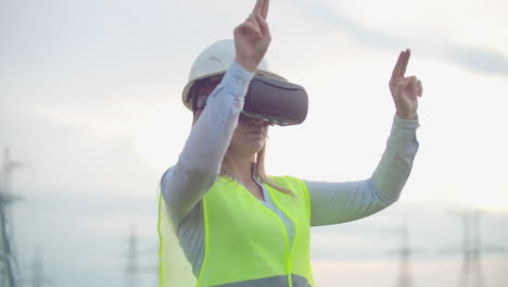 una mujer electricista con gafas de realidad virtual mueve su mano simulando el trabajo con la interfaz gráfica de una central eléctrica contra el fondo de líneas de transmisión eléctrica de alto voltaje.