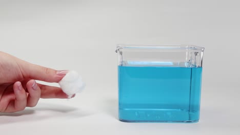 a hand places a cotton ball in a clear container filled with blue liquid, observing its buoyancy
