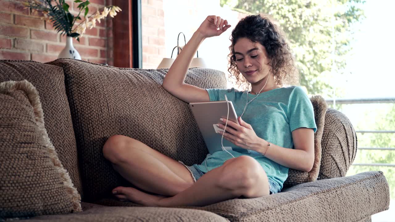 Hispanic curly hair young woman surfing the net using digital tablet and listening to music while relaxing on a couch