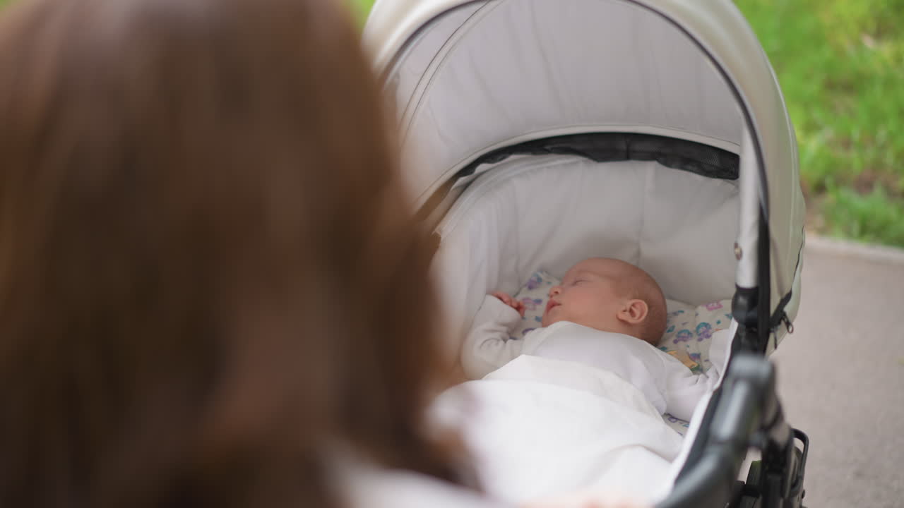 White mother watching baby in stroller, hands on handle and attentive gaze, canopy shadow and park greenery in background, protective nurturing mood, intimate lifestyle moment emphasizing bond