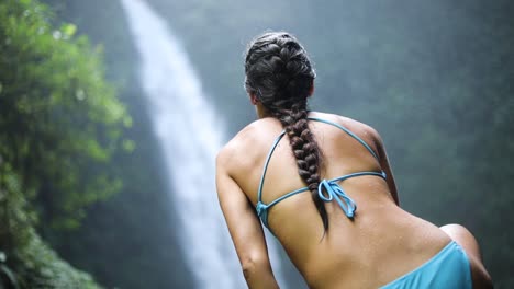 Slow-motion-panning-shot-of-a-girl-in-a-blue-bikini-sitting-in-front-of-a-gushing-NungNung-Waterfall-in-Bali,-Indonesia