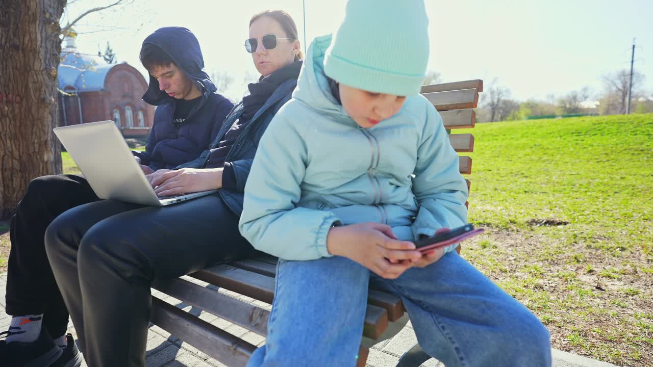 Keluarga Duduk di Bangku Taman, Sibuk dengan Laptop dan Smartphone