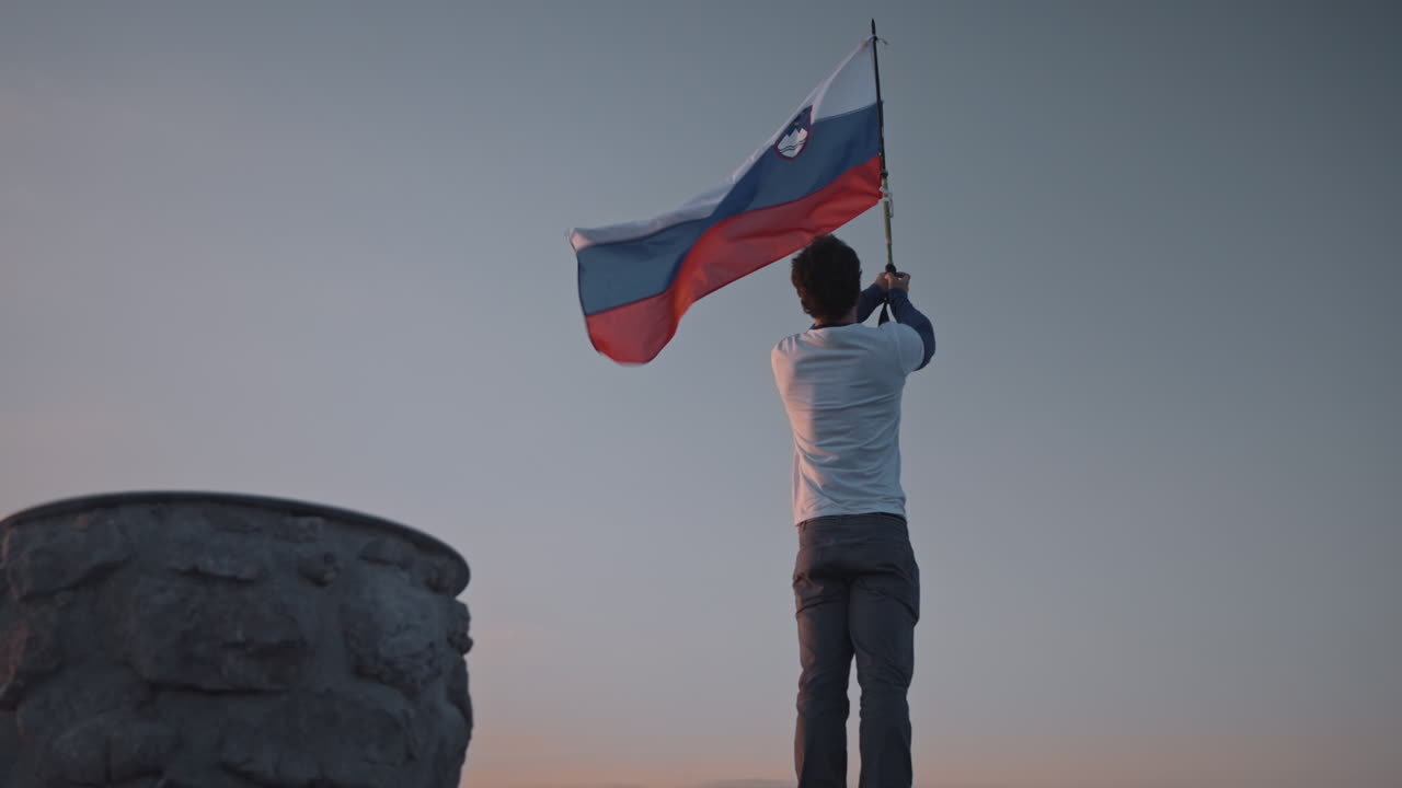 Camera circling aroud the hiker at the top of mountain Peca holding a pol with a slovenian flag attached on it, fluttering in the wind