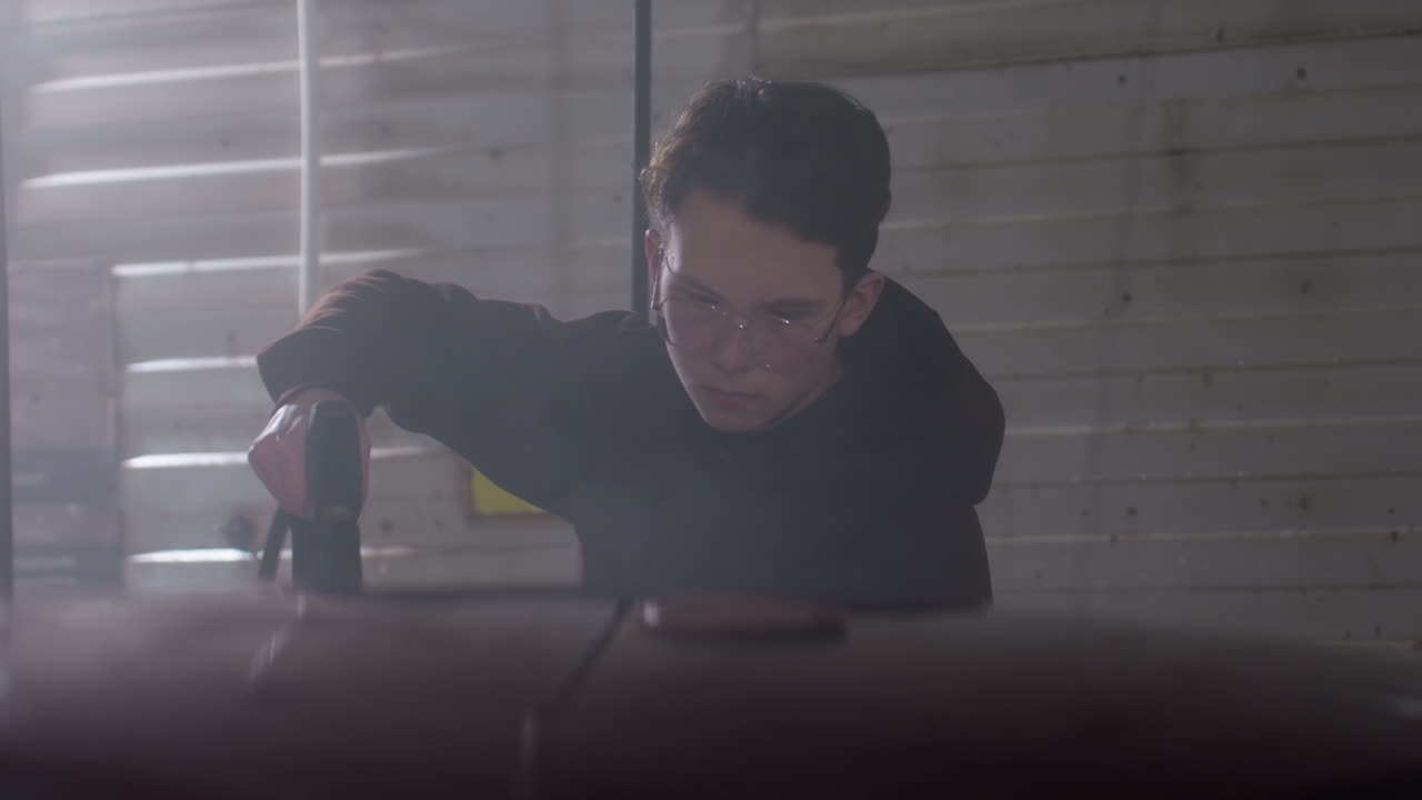 close up of red car rinse by car washer in hoodie wearing eyeglasses using high pressure hose inside dim garage capturing focused gesture under wall sign and dramatic swirling spray mist