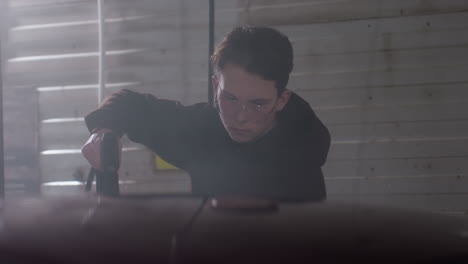 close up of red car rinse by car washer in hoodie wearing eyeglasses using high pressure hose inside dim garage capturing focused gesture under wall sign and dramatic swirling spray mist