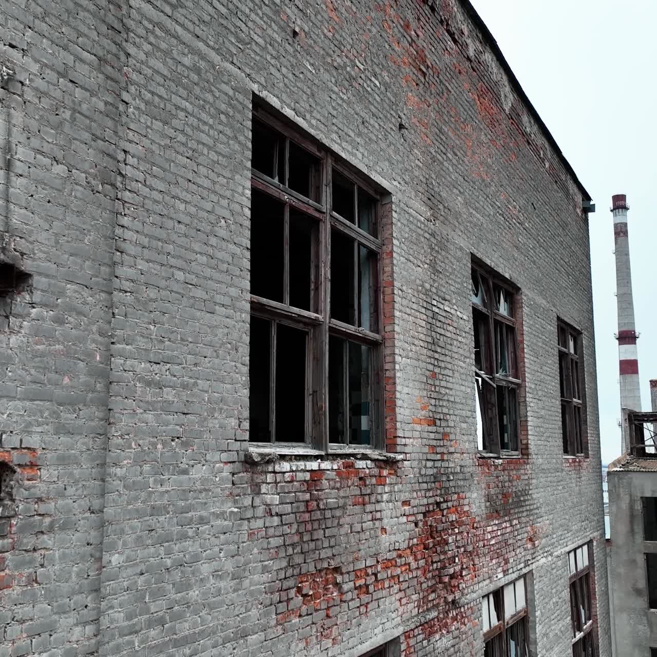 Forlorn building with crashed windows. Approaching to the brick wall of the construction. Industrial chimney pipe at the backdrop