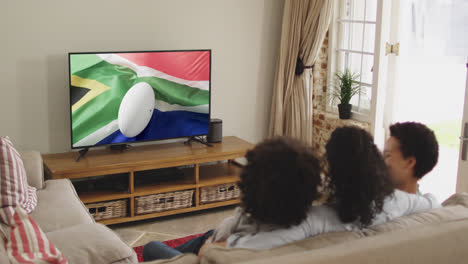 familia biracial viendo la televisión con una pelota de rugby con la bandera de sudáfrica en la pantalla