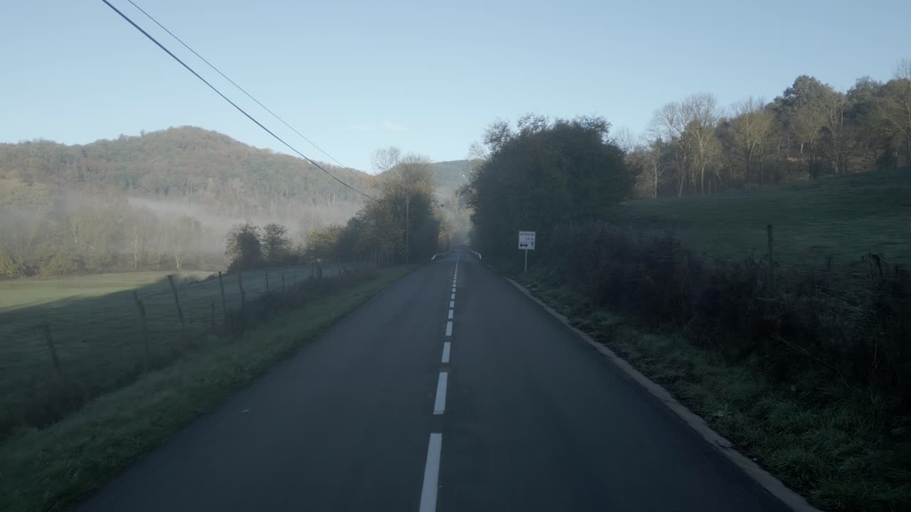 Morning fog lingers over a narrow asphalt road winding through the green hills of a mediterranean landscape, creating a serene and picturesque scene at sunrise
