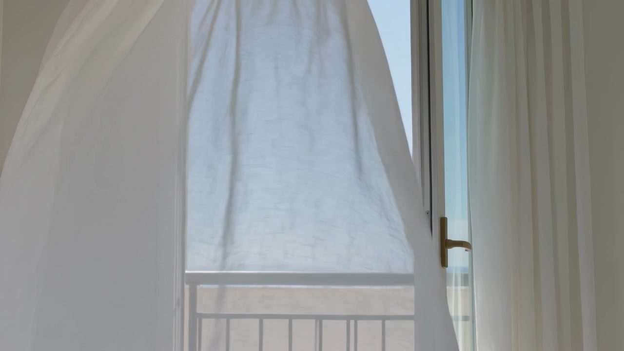 Brezza che solleva una tenda trasparente che ondeggia attraverso la porta del balcone a casa, con la luce solare che illumina le pieghe sottili.