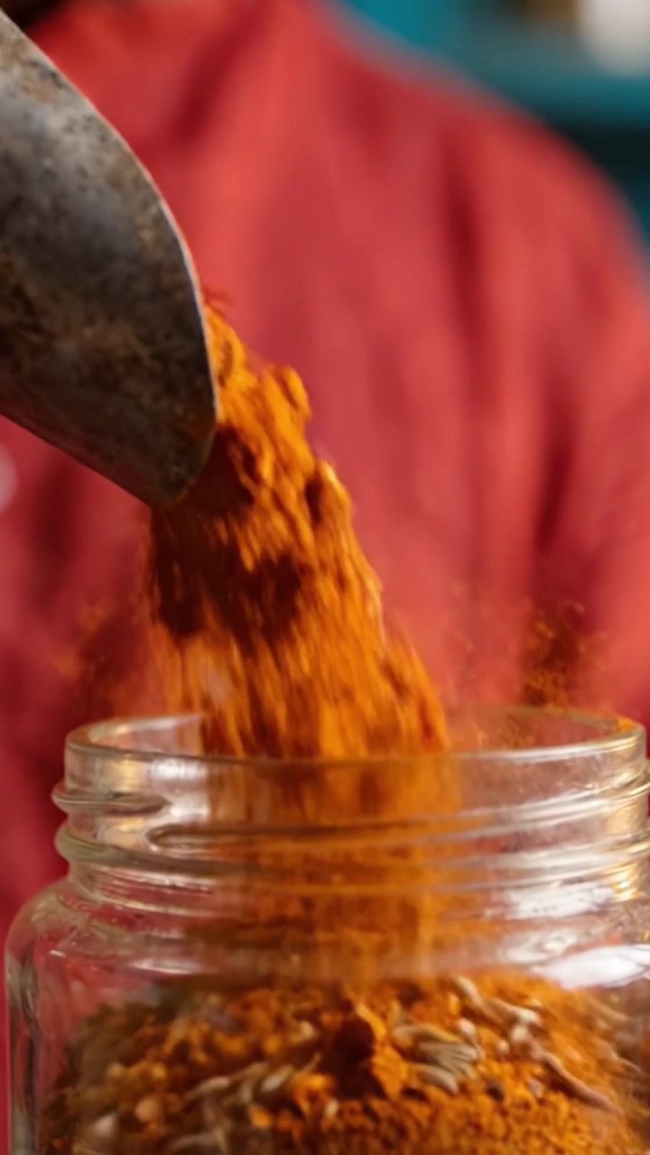 Una persona midiendo y preparando especias aromáticas con una cuchara, destacando cúrcuma vibrante y semillas fragantes en un frasco de vidrio, capturando la esencia de la cocina