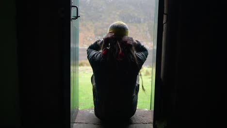 a man with dreadlocks and wearing hiking gear walks into shot and sits on a step in a narrow doorway of a bothy in the highlands of scotland to enjoy the view outdoors