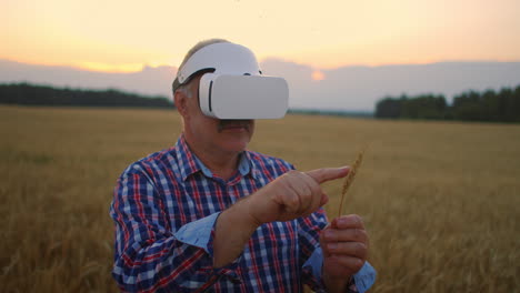 Senior-adult-farmer-in-a-virtual-reality-helmet-in-a-field-of-grain-crops.-In-the-sunset-light-an-elderly-man-in-a-tractor-driver-uses-virtual-reality-glasses.-VR-technologies-and-modern-agribusiness