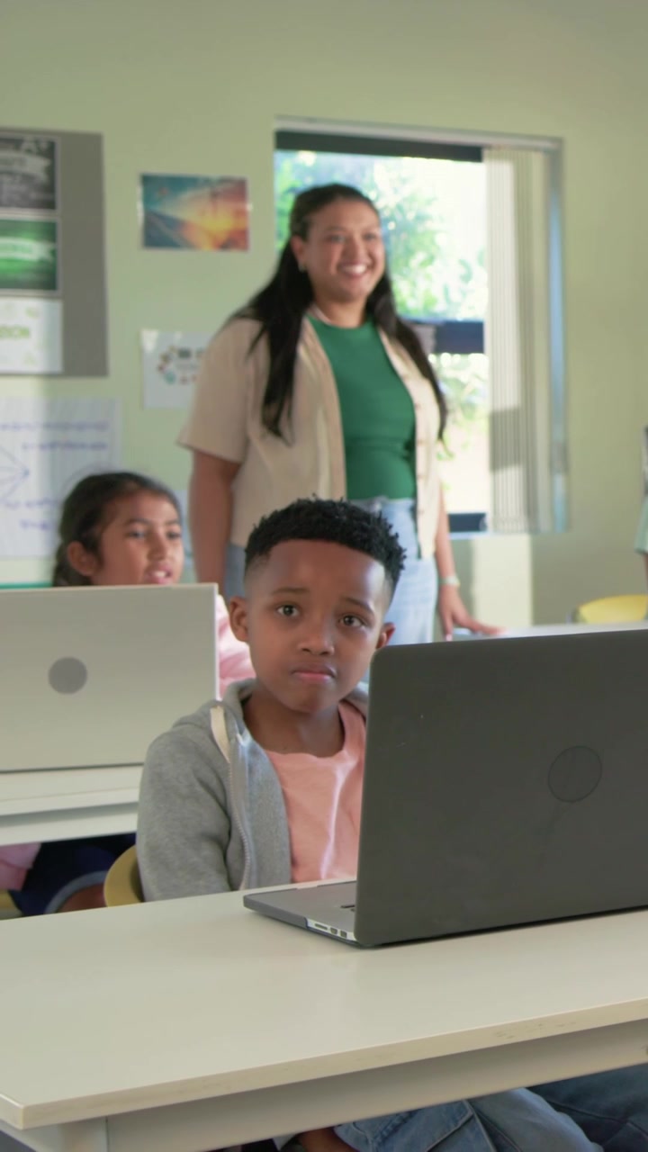 Vertical video: Smiling teacher in green top guiding boy looking at laptop in class with tablet