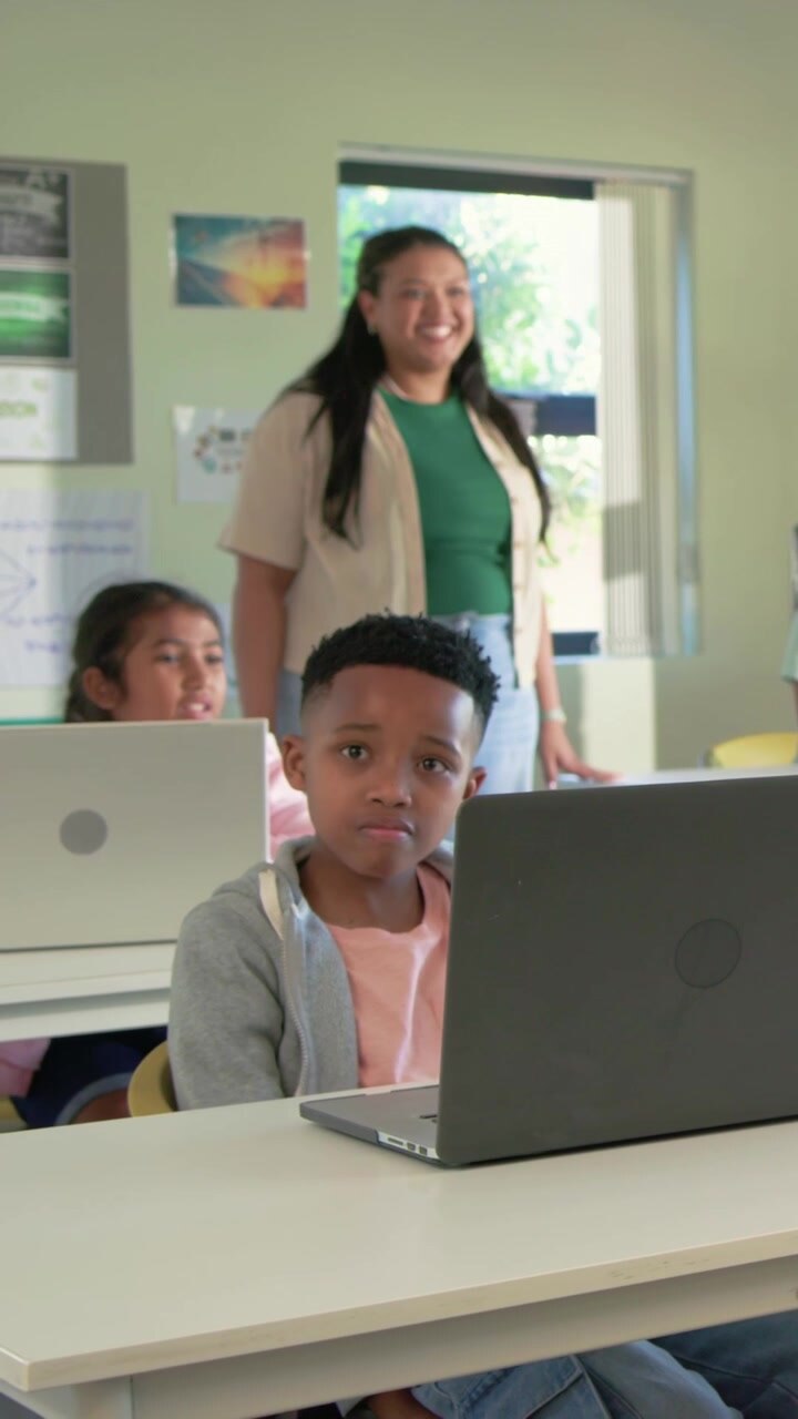 Vertical video: Smiling teacher in green top guiding boy looking at laptop in class with tablet