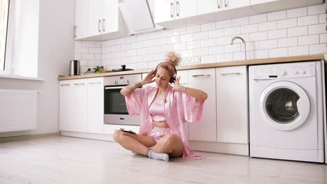 joven feliz con cabello rosa sentada en el suelo de la cocina con pijama rosa y escuchando música con auriculares