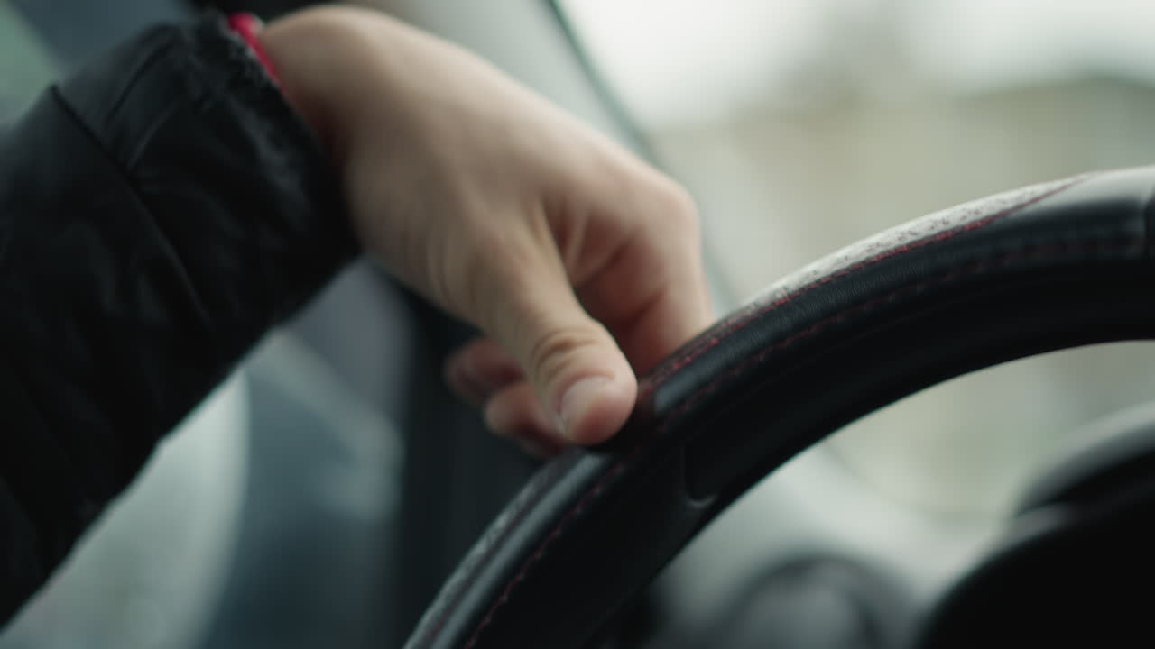 Young man hands gripping leather steering wheel with red stitching, fingers resting on rim while guiding car through wet street, showing driver focus, road view through side window blurred in motion