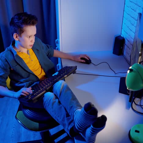 teen boy is enjoying computer games at home. boy playing computer video games while sitting in the chair with his legs on desk. top view.