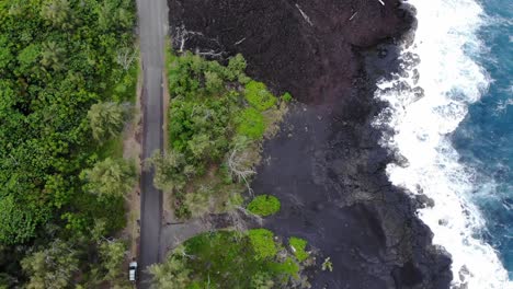 Flight-along-volcanic-coastline-of-Hawaii-with-a-road,-parked-car,-black-rock-beach-and-trees